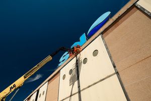 Worker on yellow crane lift installs large blue circular sign on beige building facade against blue sky.