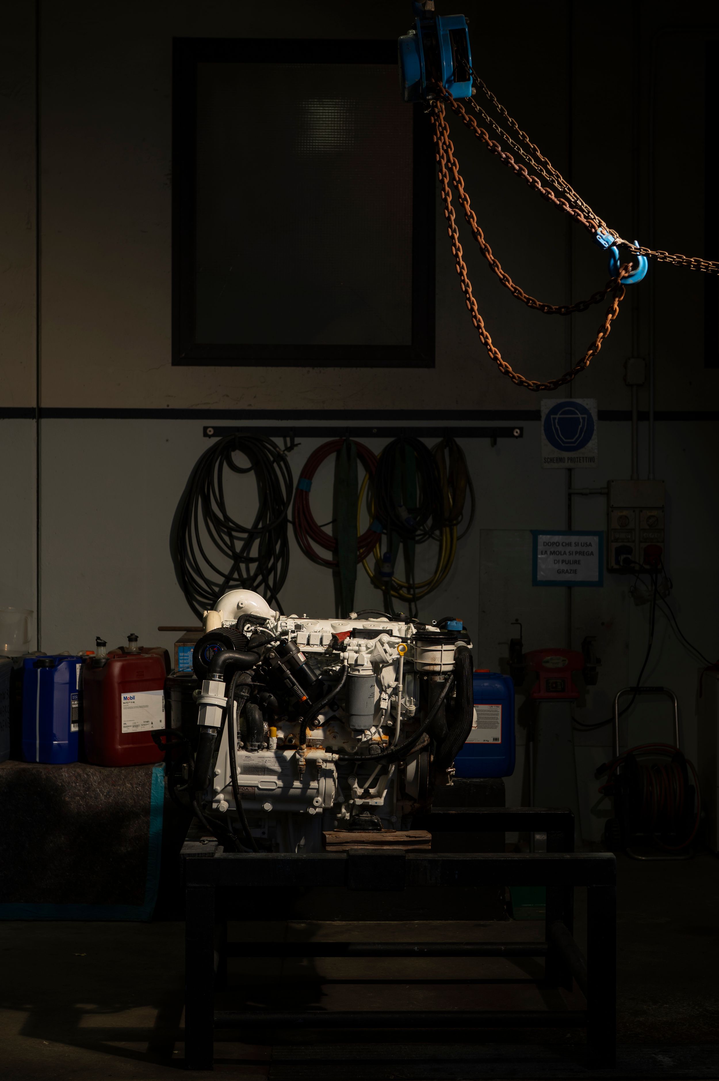 Engine on workbench in dark garage with orange chains, hoses, and equipment on wall.