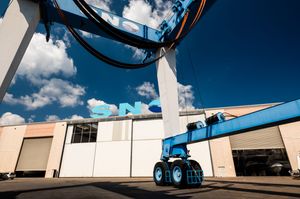 Blue industrial crane machine outdoors near a warehouse building under a partly cloudy sky.