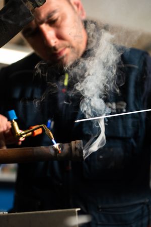 A mechanic uses a blowtorch to solder a copper pipe, with smoke rising in a workshop.