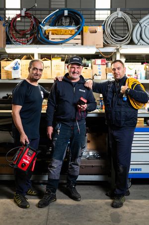 Three male technicians pose together in a well-equipped workshop holding tools and equipment.