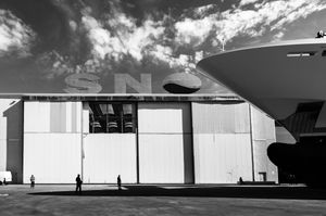 Black and white photo of a large ship docked beside a massive industrial hangar with partial letters visible.