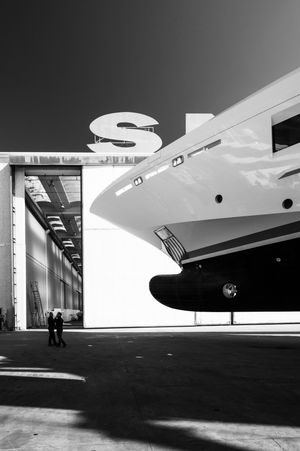 Black and white photo of a large yacht being built inside a shipyard hangar with two workers standing nearby.