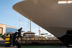 A scuba diver in full gear inspects the hull of a large white yacht docked at a marina on a clear day.