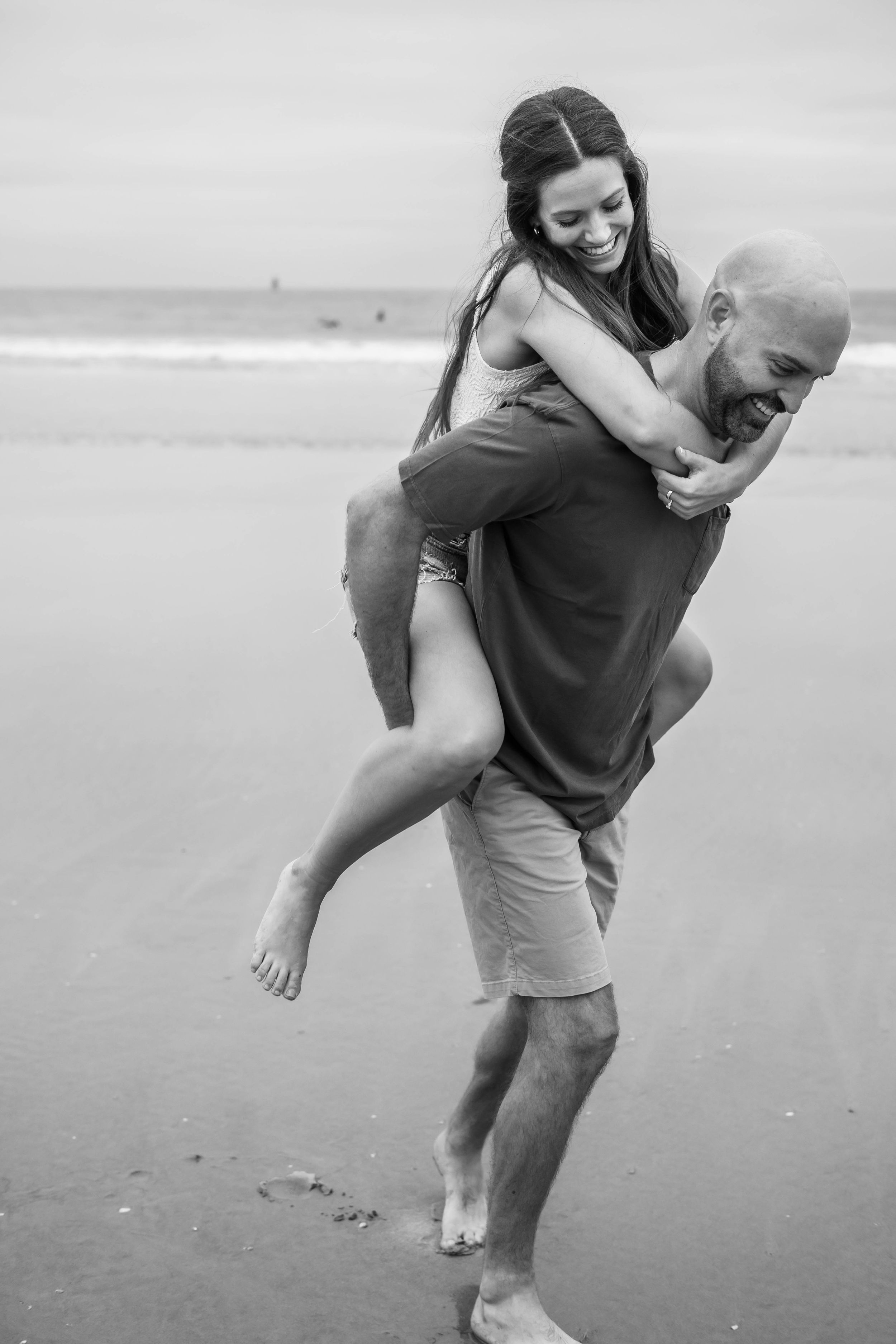 A couple shares a playful moment on the beach as they embrace in a black and white photograph.