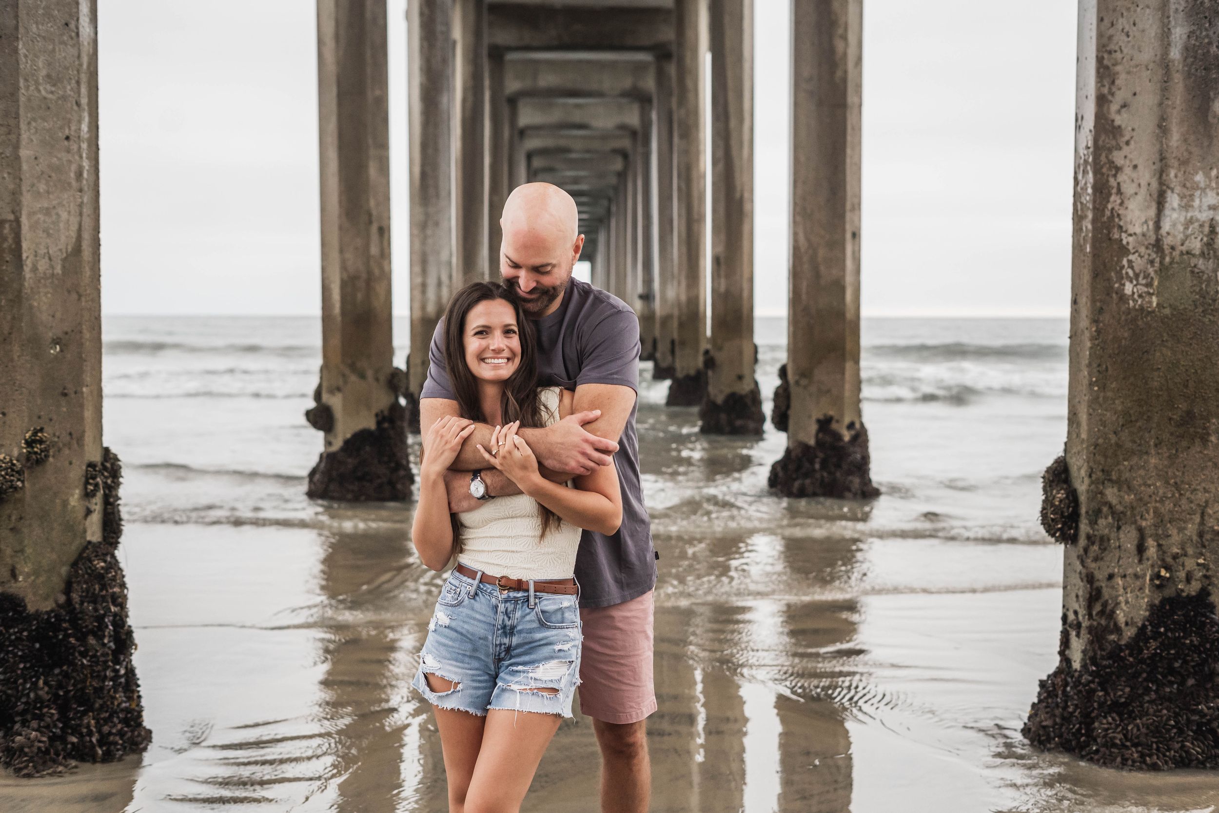 A couple shares a romantic moment under a wooden pier at the beach during low tide.