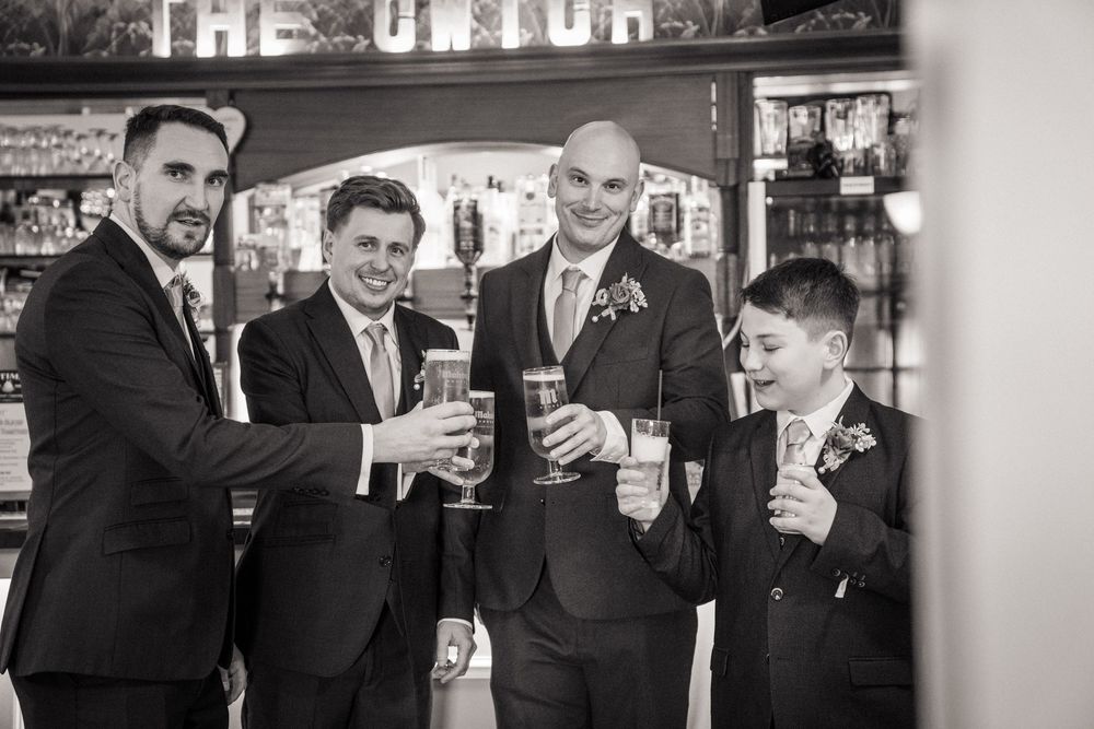A black and white photo of groomsmen toasting with drinks at a bar during a wedding celebration.