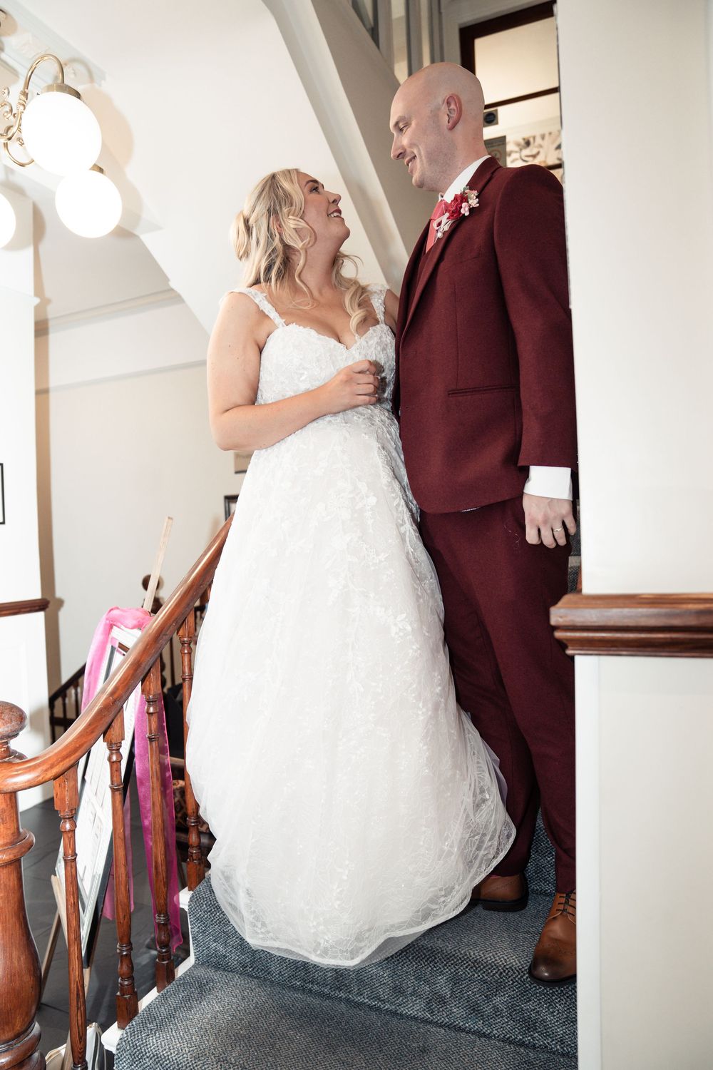 A couple shares a joyful moment on a staircase, with the bride in white lace and groom in burgundy suit.