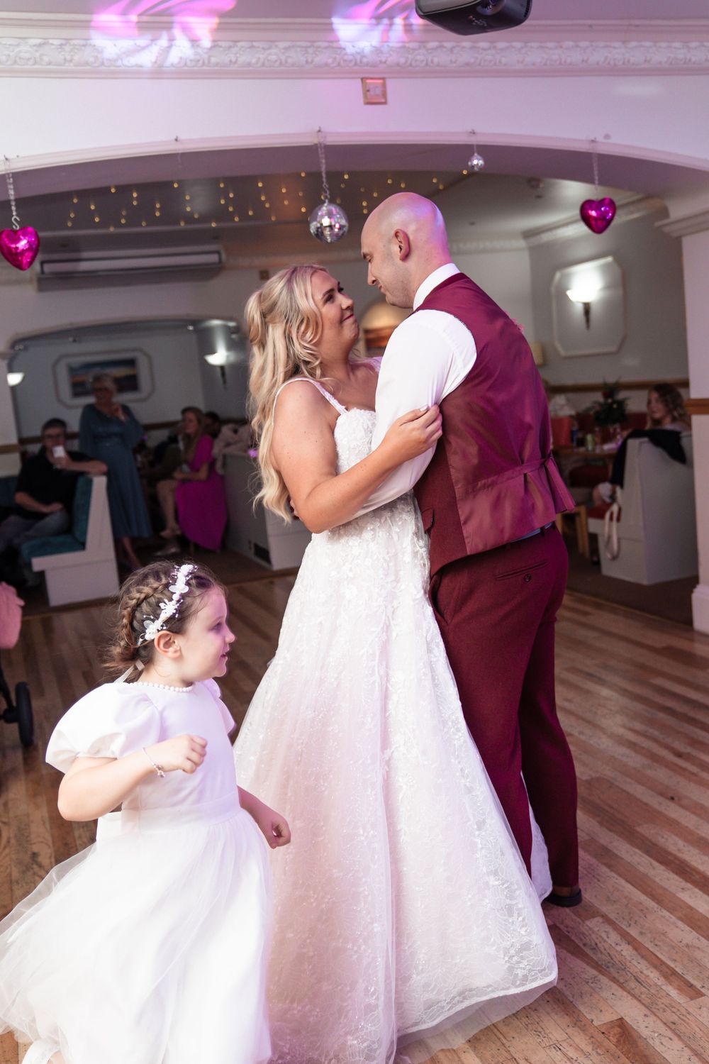 A couple shares their first dance at a wedding reception while a young girl in white watches from the side.