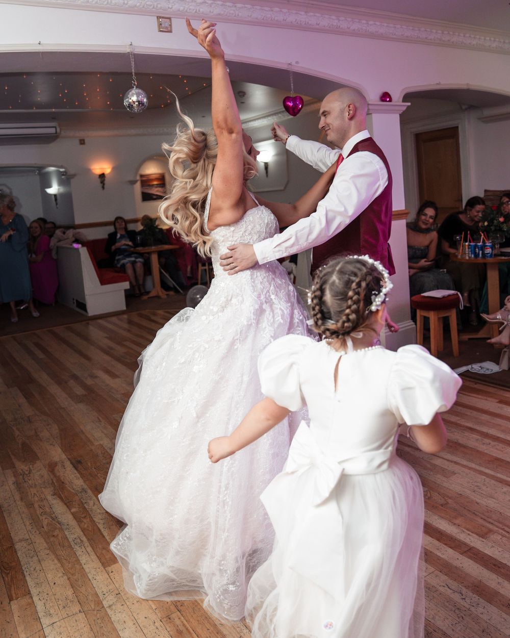A couple dances joyfully at their wedding reception on a wooden dance floor while a flower girl in white watches.