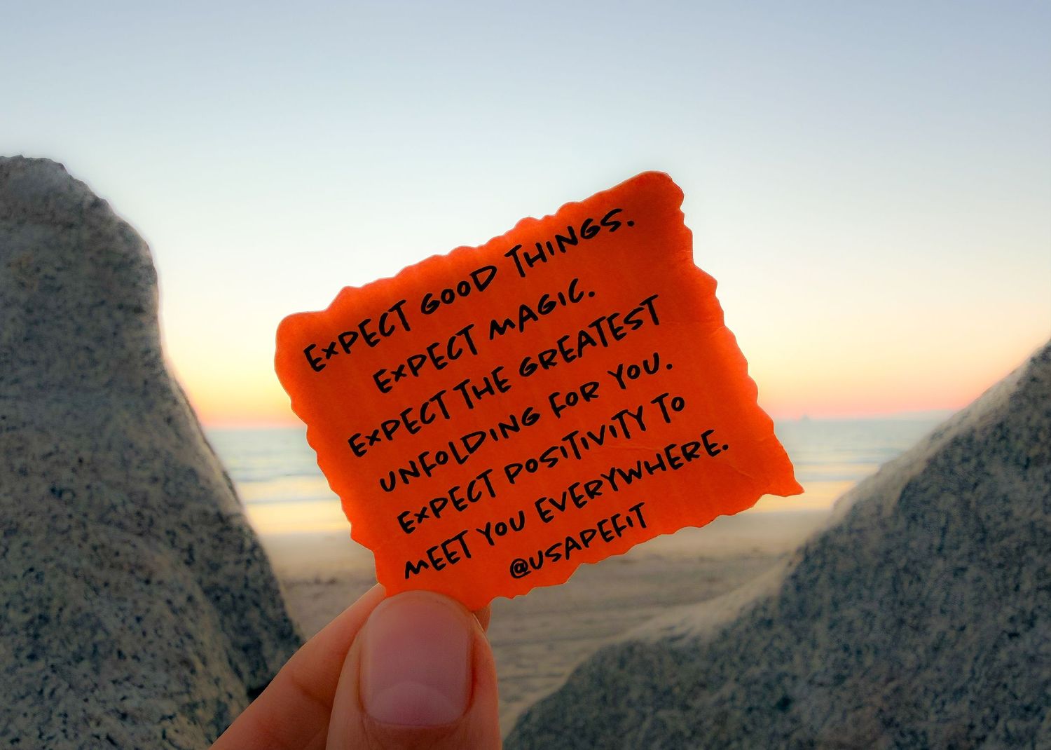 Hand holding an orange sticky note with text against a blurry coastal sunset background.