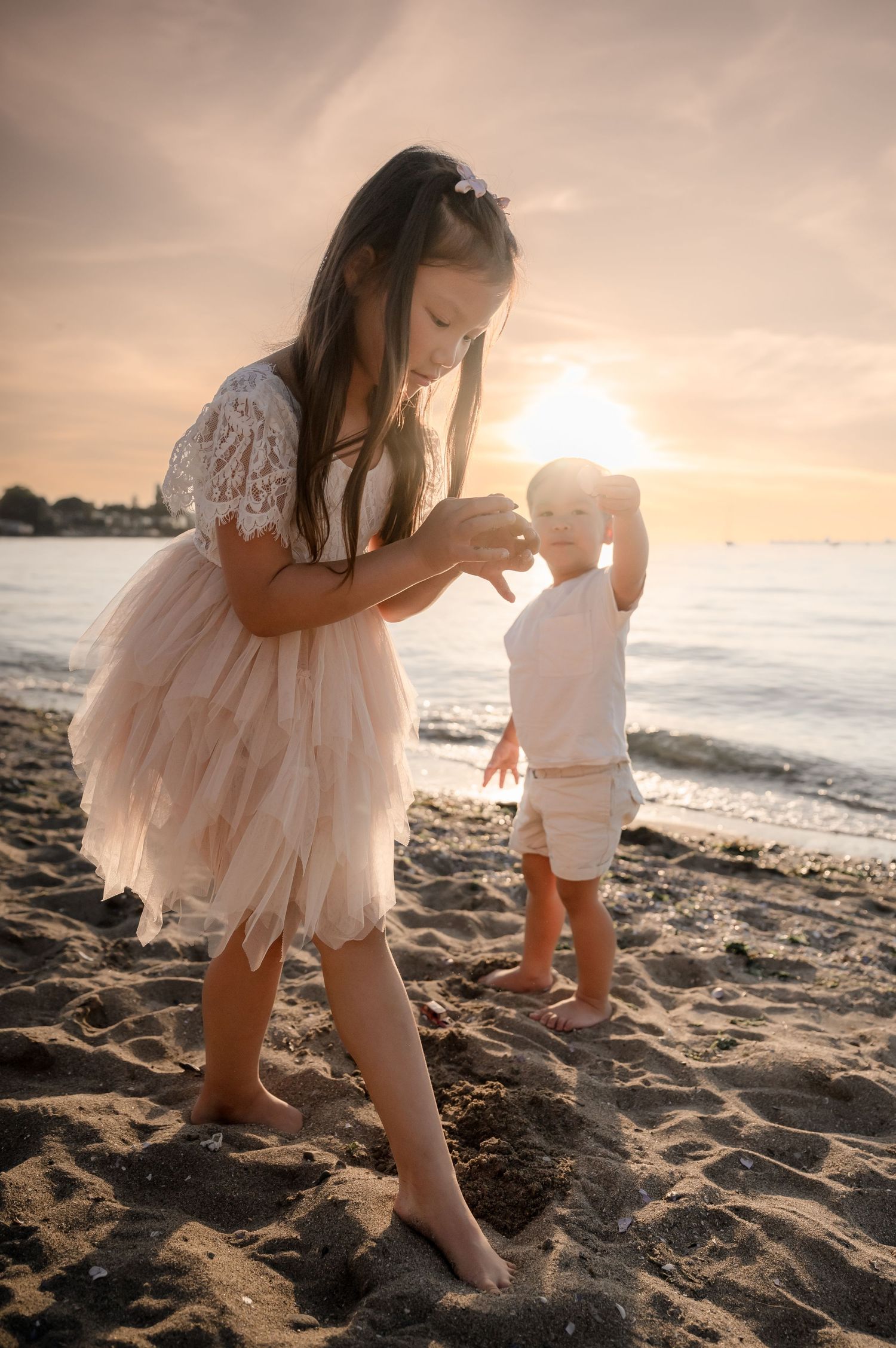 Two children in white outfits play together on a sandy beach at sunset creating a dreamy coastal scene.