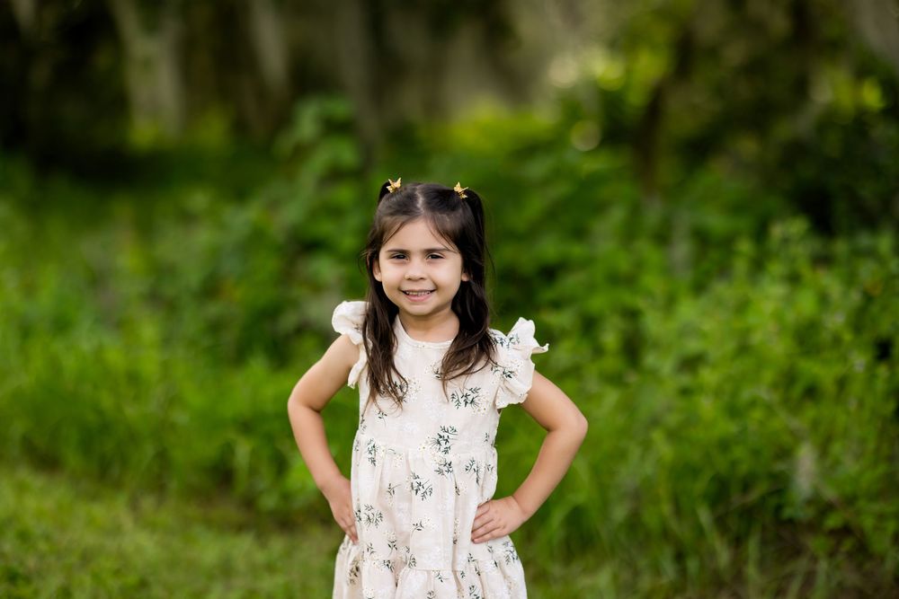 Young child in a white floral dress poses with hands on hips in a lush green outdoor setting.
