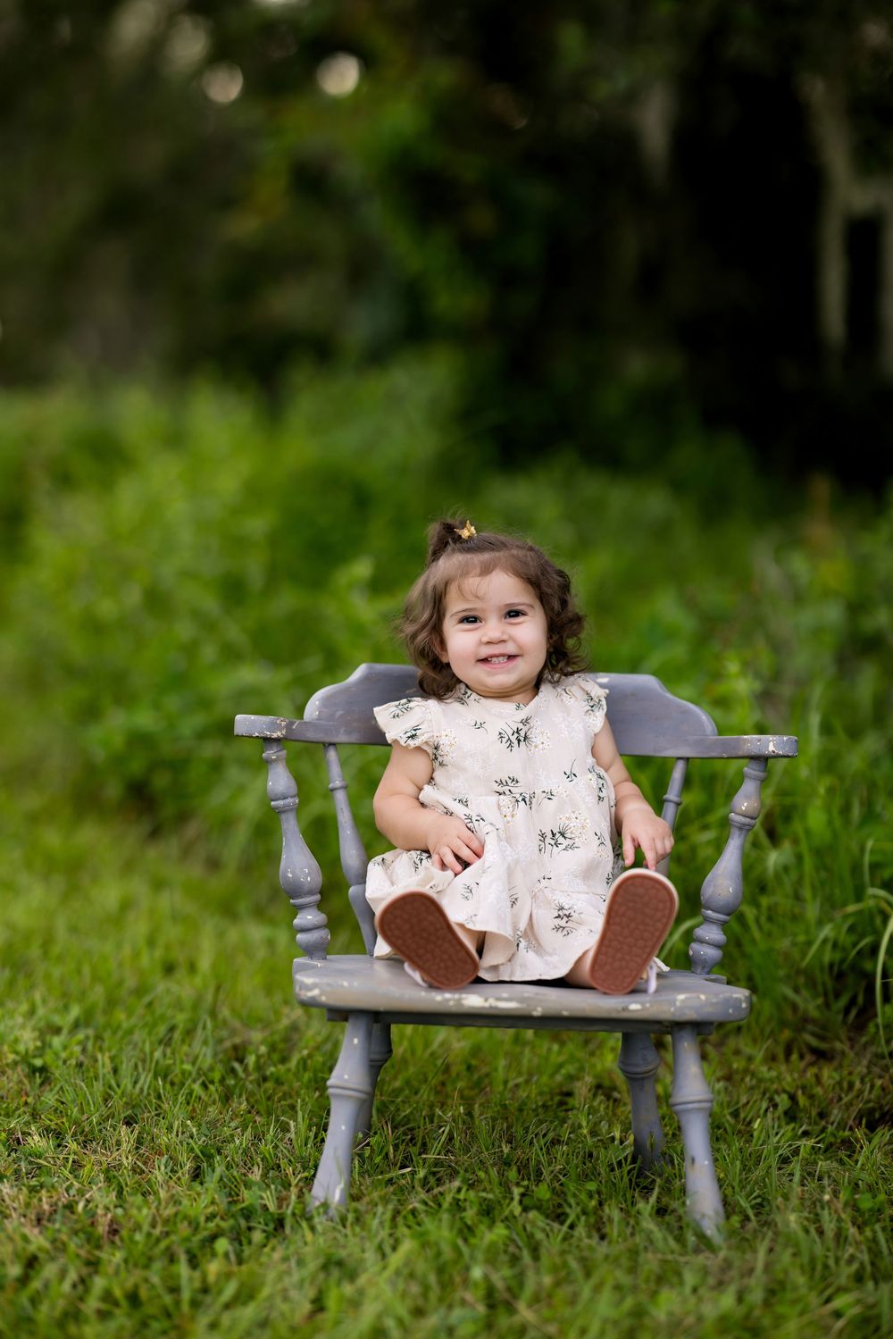 A child sits on a rustic gray wooden bench surrounded by lush green grass in an outdoor setting.