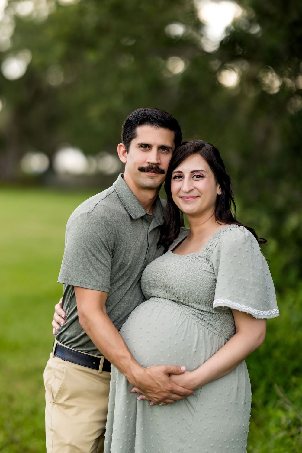 A couple embraces during a maternity photo shoot in a lush green outdoor setting.