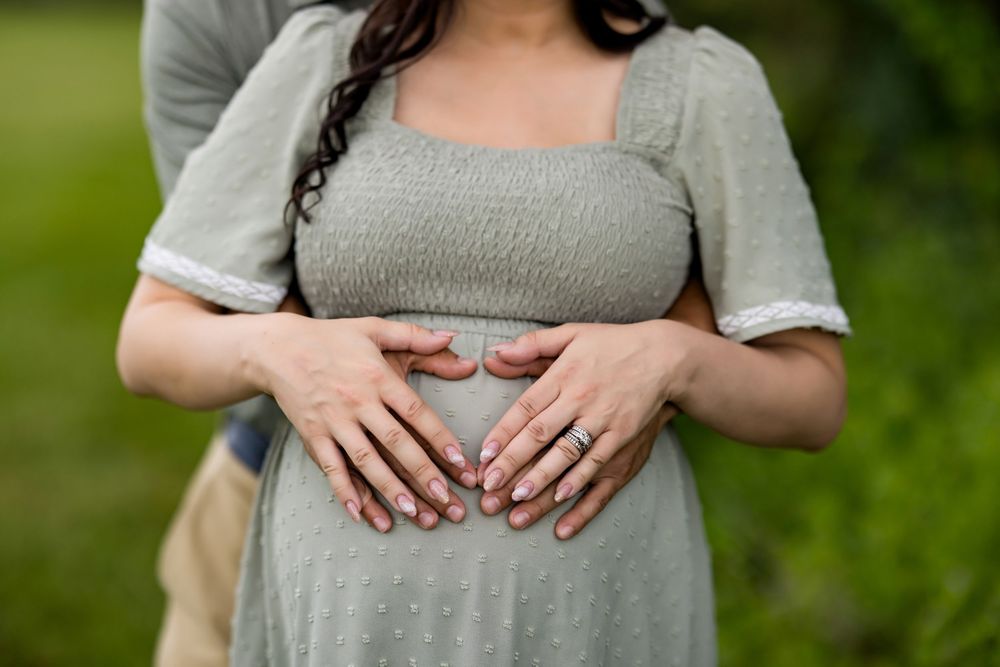 Hands form a heart shape over a pregnant belly in a grey dress against a blurred green outdoor background.