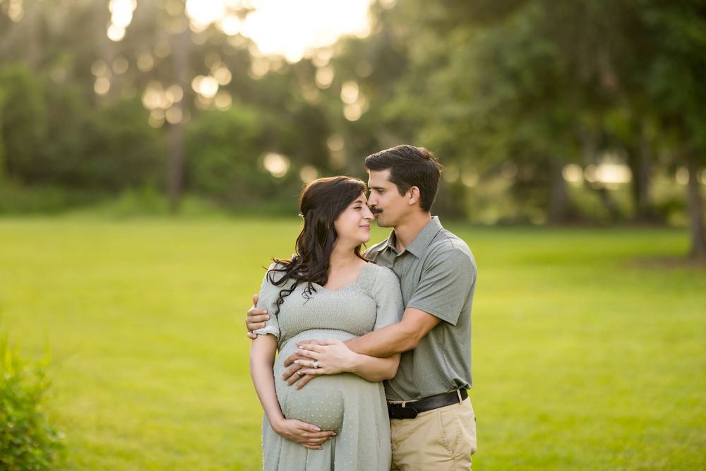 A couple embraces in a sunny green field during a maternity photoshoot at golden hour.