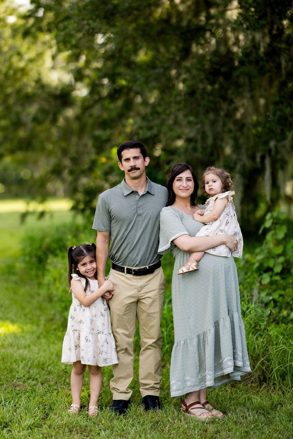 A family poses together in a lush green outdoor setting wearing coordinated casual outfits in neutral tones.