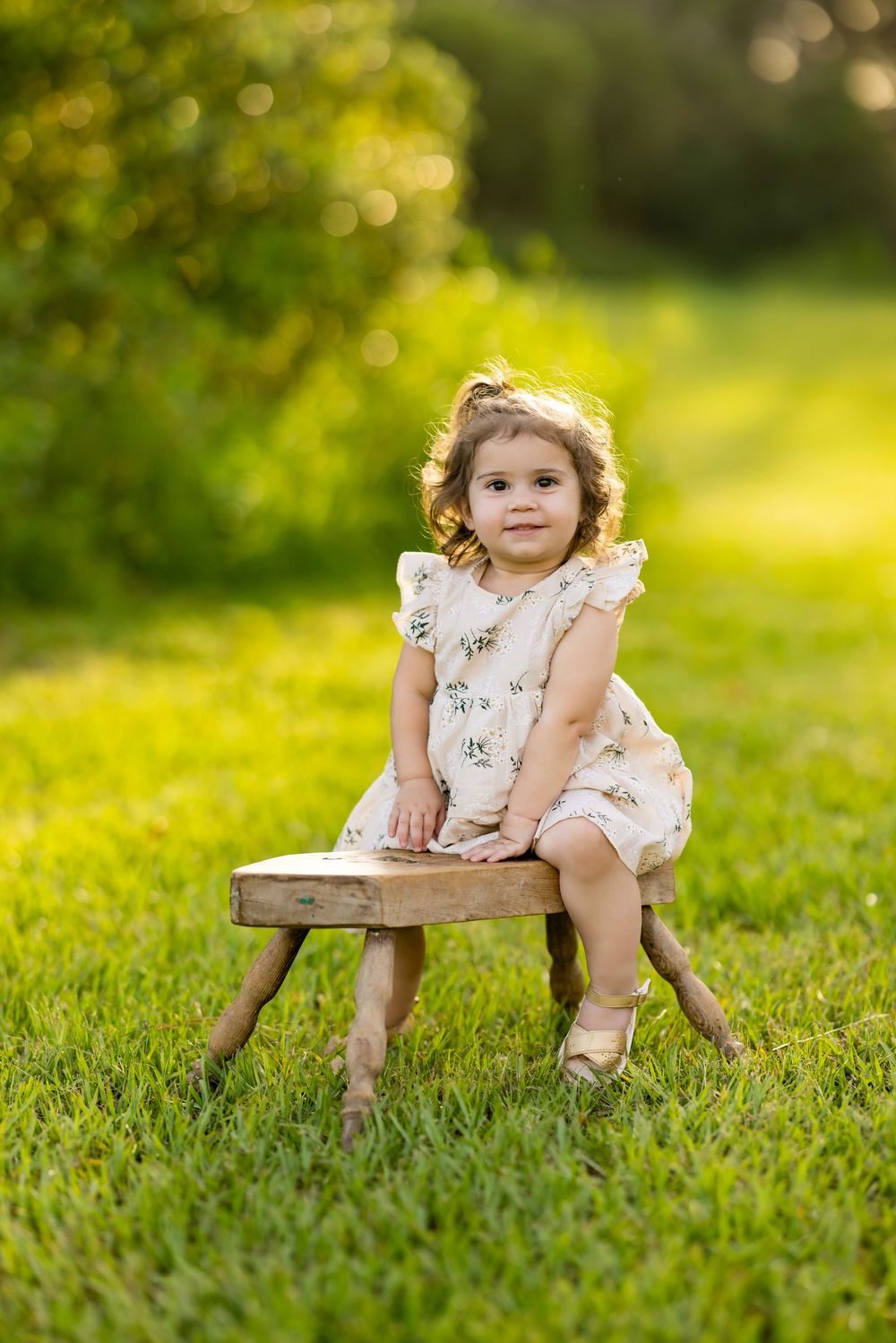 A child in a white dress sits on a wooden chair in a sunlit grassy field with bokeh effect in the background.