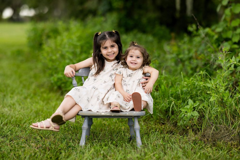 Two children sit together on a wooden bench in a lush green garden setting.
