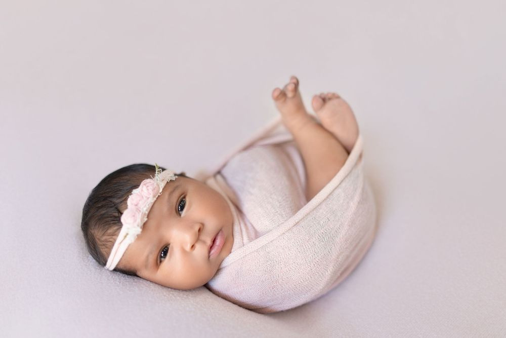 A baby wearing a white headband and wrap lies on a light gray background while stretching their leg.