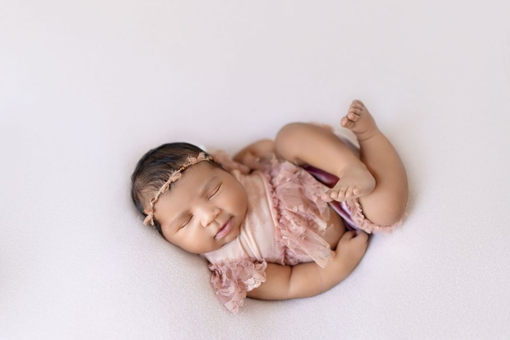 Sleeping newborn baby wearing pink lace dress curled up in fetal position on white blanket during professional photoshoot.