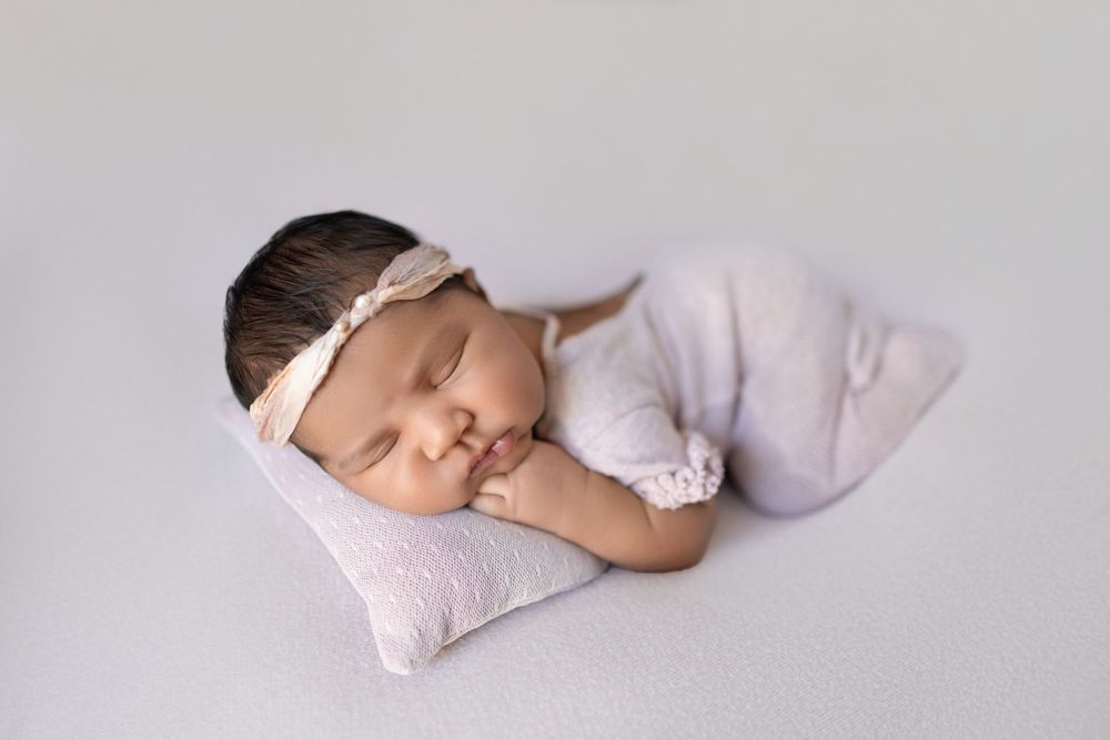 A sleeping newborn baby wearing a light pink outfit lies peacefully on a soft blanket in a professional studio setting.