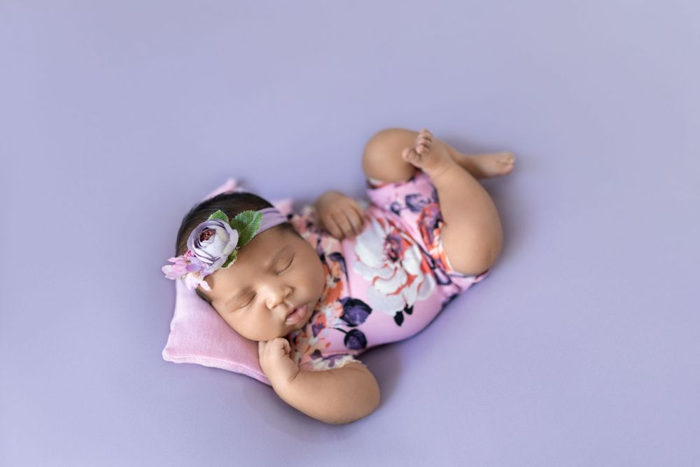 Newborn photography session with delicate pink flowers and soft purple backdrop creating a dreamy atmosphere.