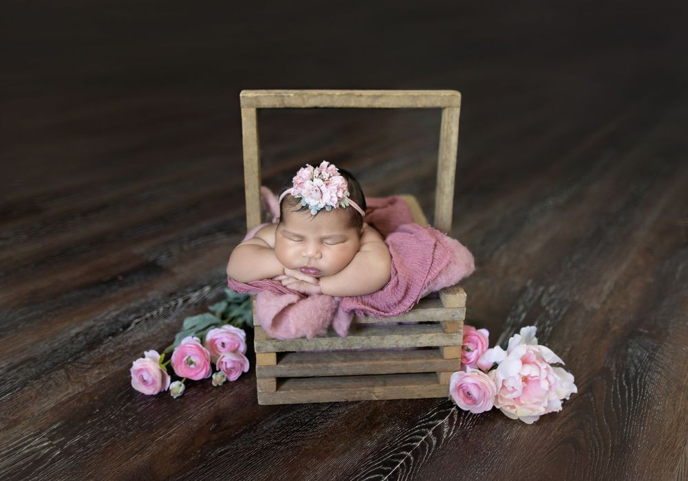 Artistic newborn portrait with rustic wooden frame prop and scattered pink rose petals on dark wood floor.