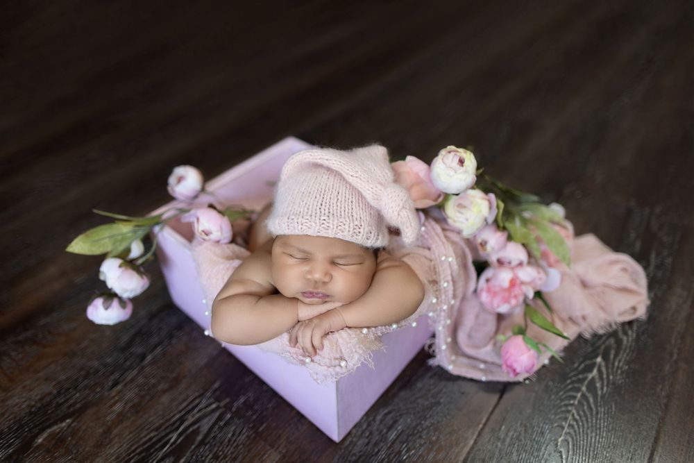 Newborn baby sleeps peacefully on wooden surface surrounded by delicate pink flowers and soft fabrics.