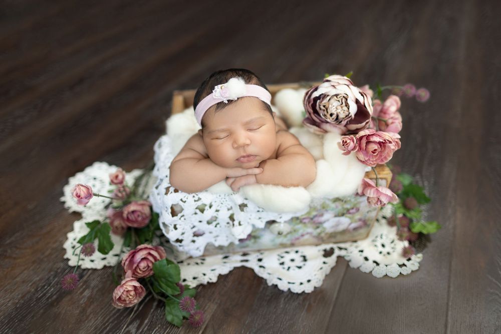 Sweet newborn photography setup with vintage lace doily and pink floral decorations on dark wooden surface.