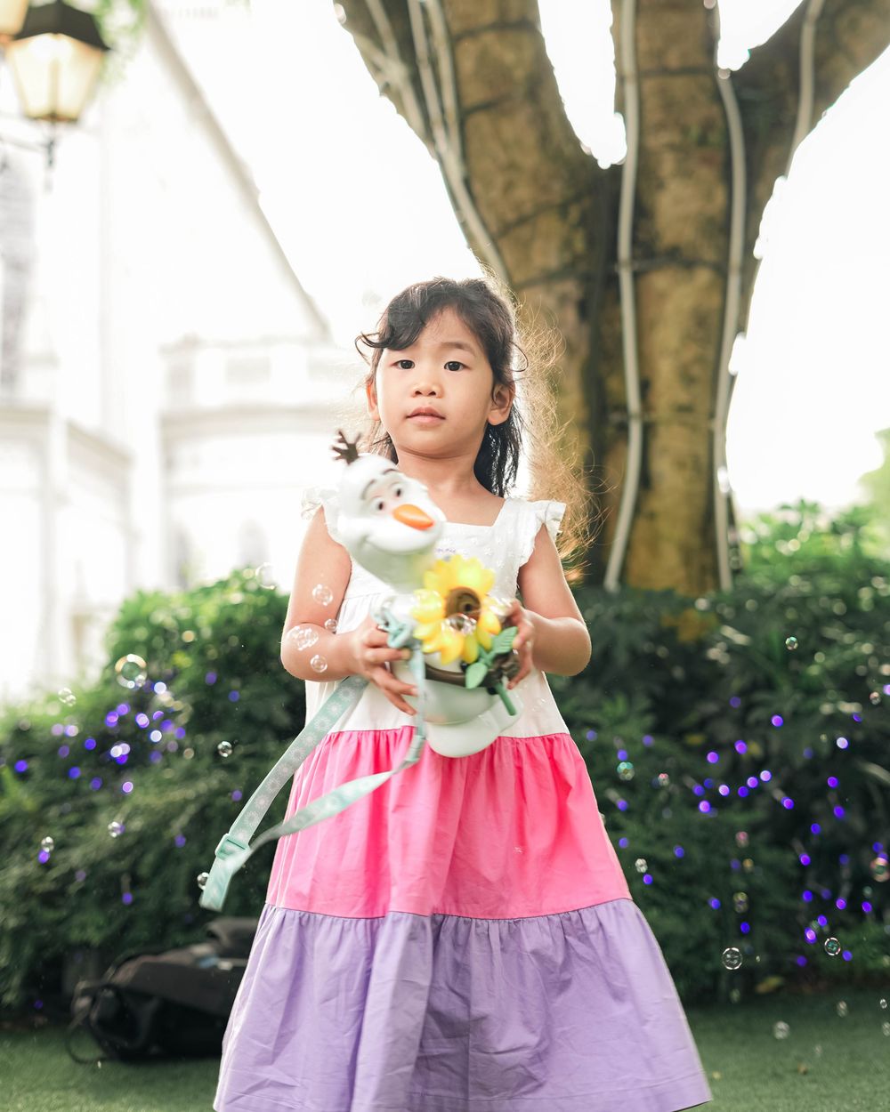 Young child in a pink and purple dress poses playfully in a garden setting with sunflowers and purple flowers.