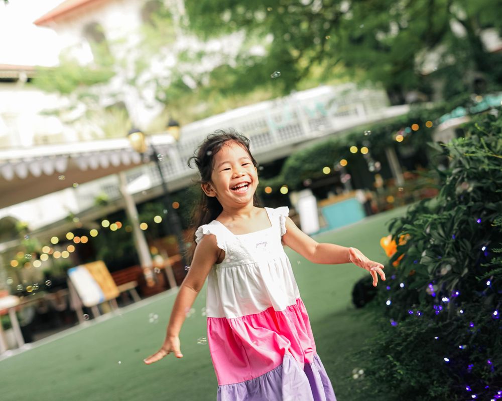 A child in a white and pink dress dances joyfully on a green lawn with string lights and garden scenery in the background.