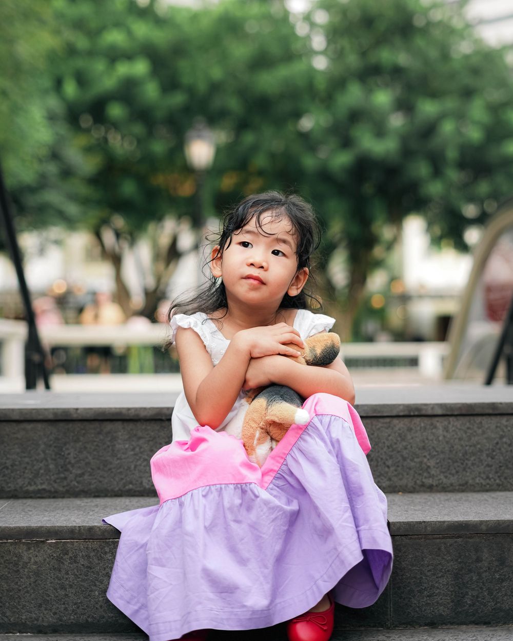 A child in a purple and pink dress sits thoughtfully on a stone ledge in a park setting.