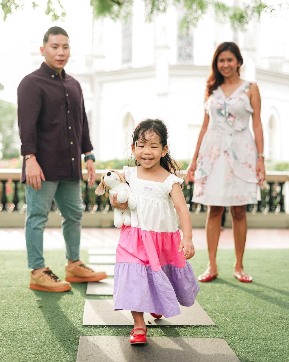 Children play and walk together on green turf at an outdoor venue wearing colorful summer dresses and casual attire.