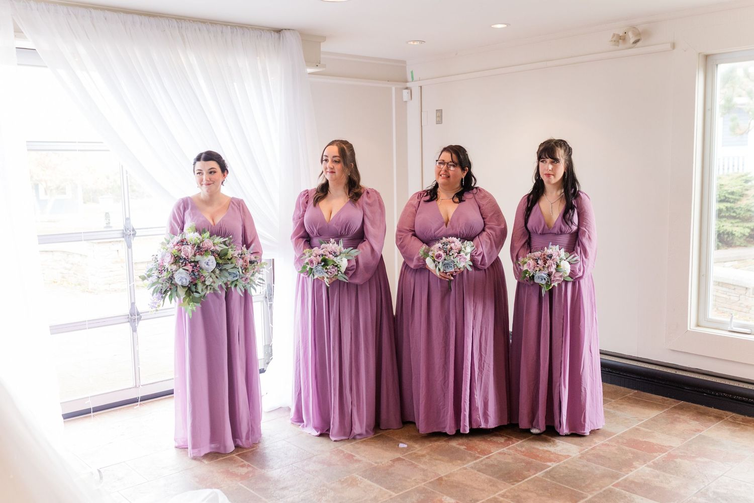 Bridesmaids in matching dusty purple dresses stand together in a bright white room during a wedding celebration.