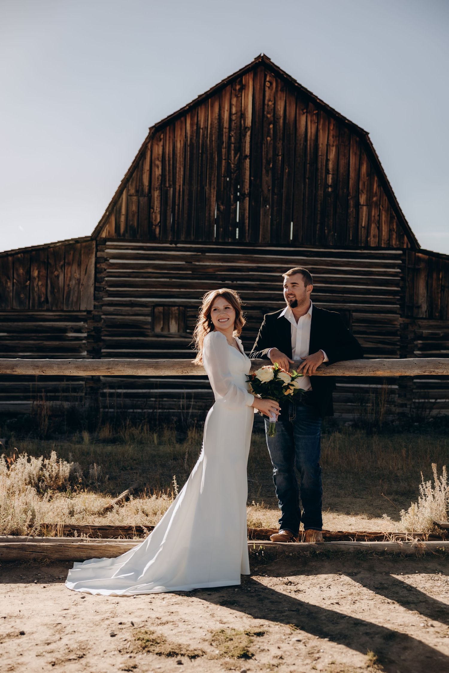 A couple dances joyfully in front of a rustic wooden barn on their wedding day in golden sunlight.