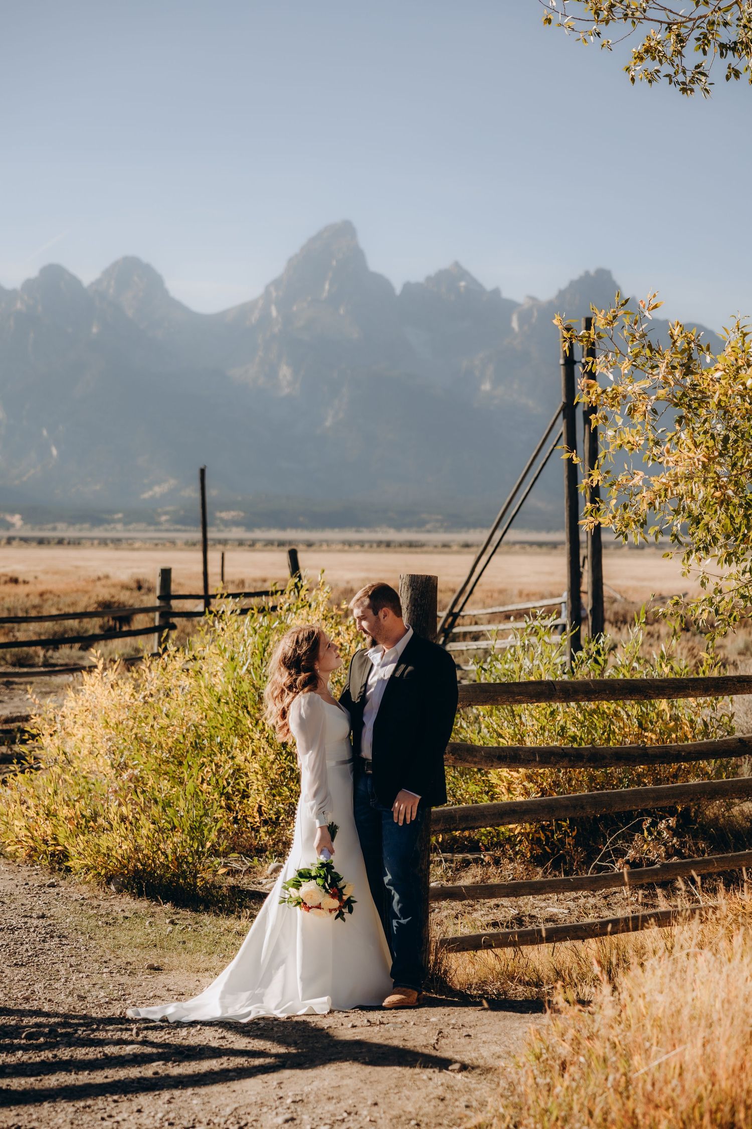 A couple sits on a rustic wooden fence with majestic mountain peaks rising in the background during golden hour.