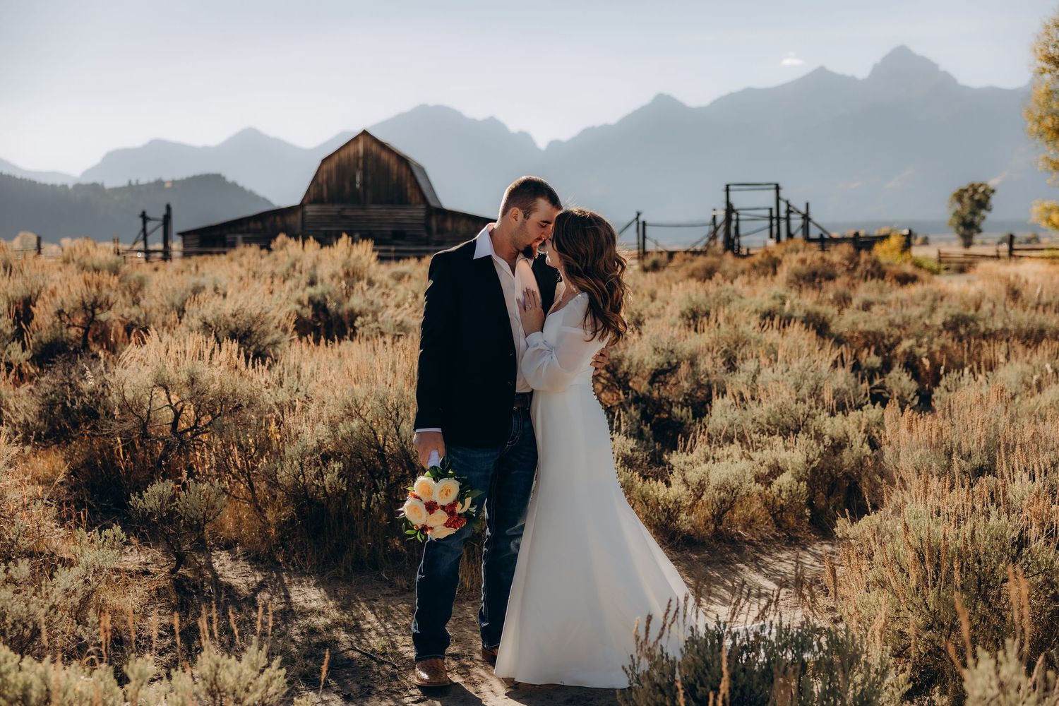 A couple shares an intimate moment against a backdrop of majestic mountain peaks and golden desert brush at sunset.
