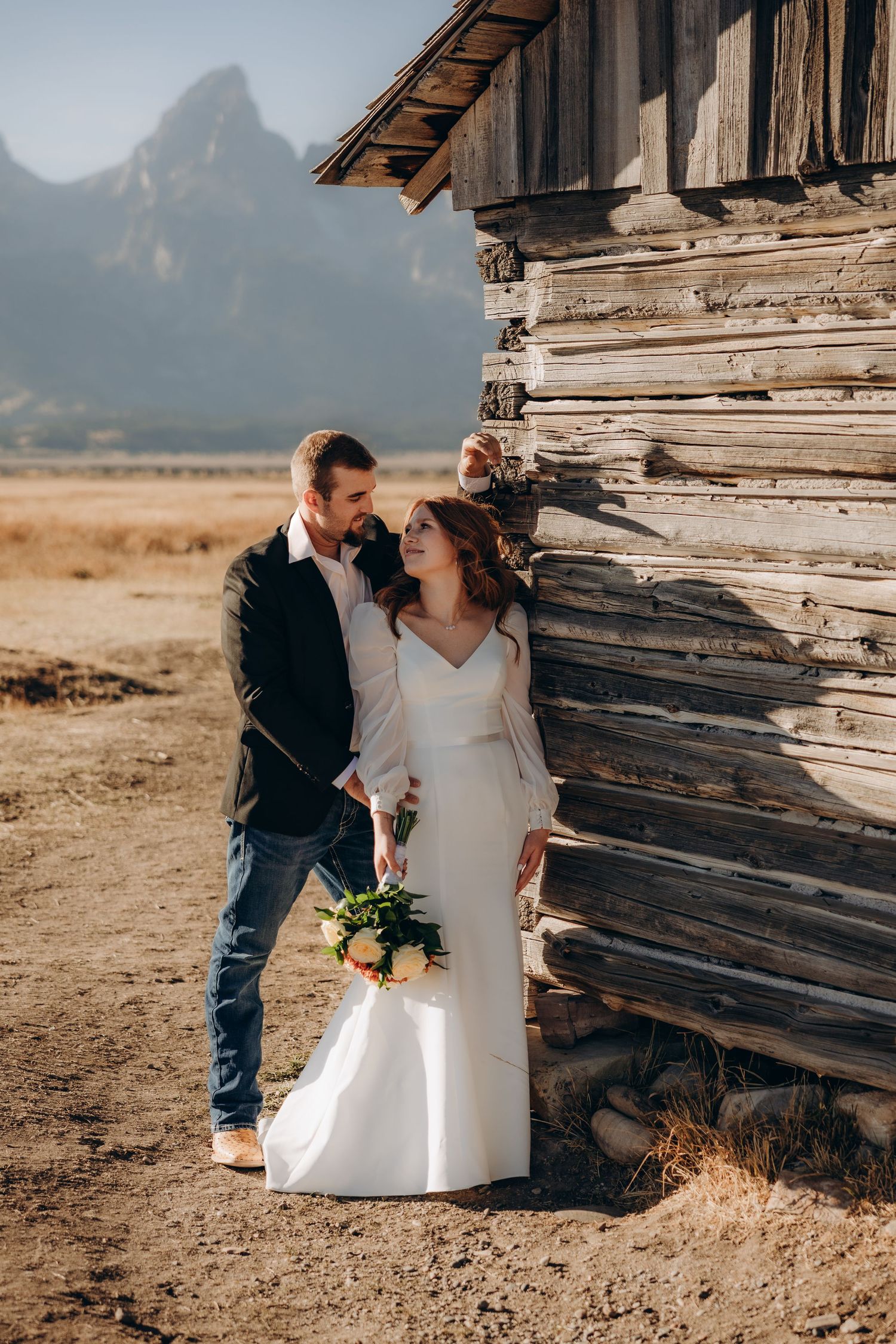 Romantic wedding couple poses against rustic wooden backdrop in scenic mountain landscape.