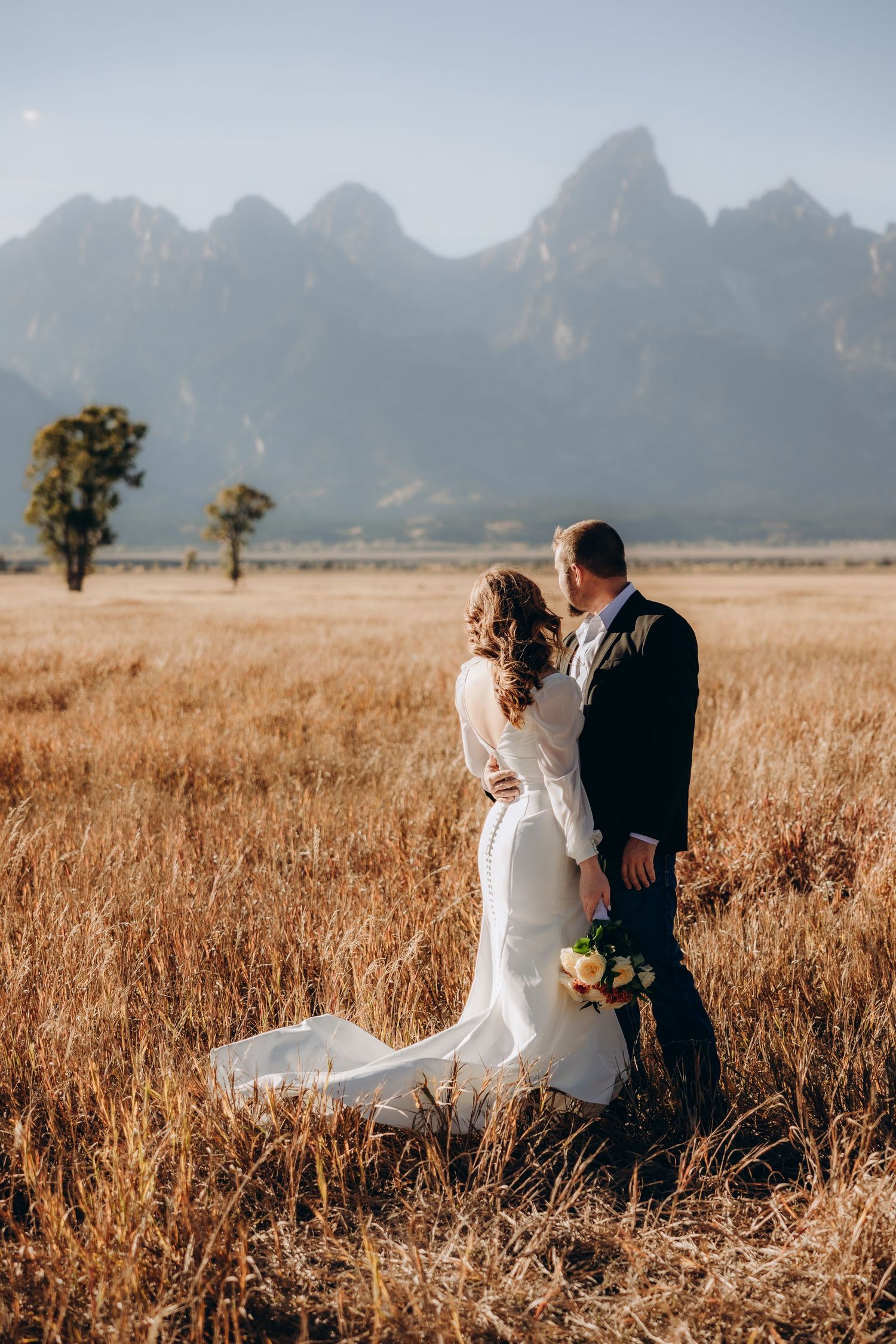 A romantic wedding photoshoot in a golden field with majestic mountains in the background at sunset.