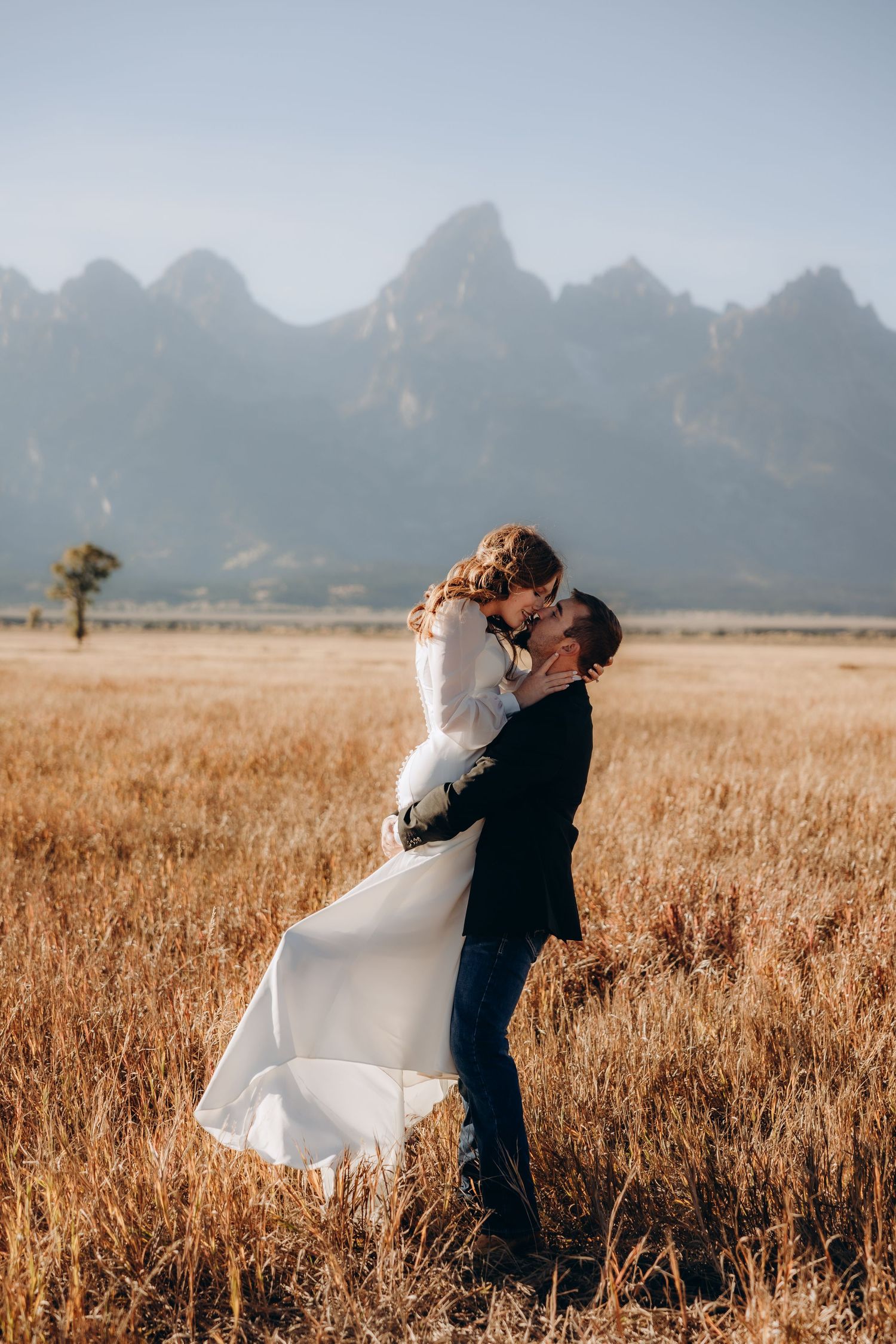 A couple embraces in a golden wheat field with majestic mountain peaks rising in the misty background.