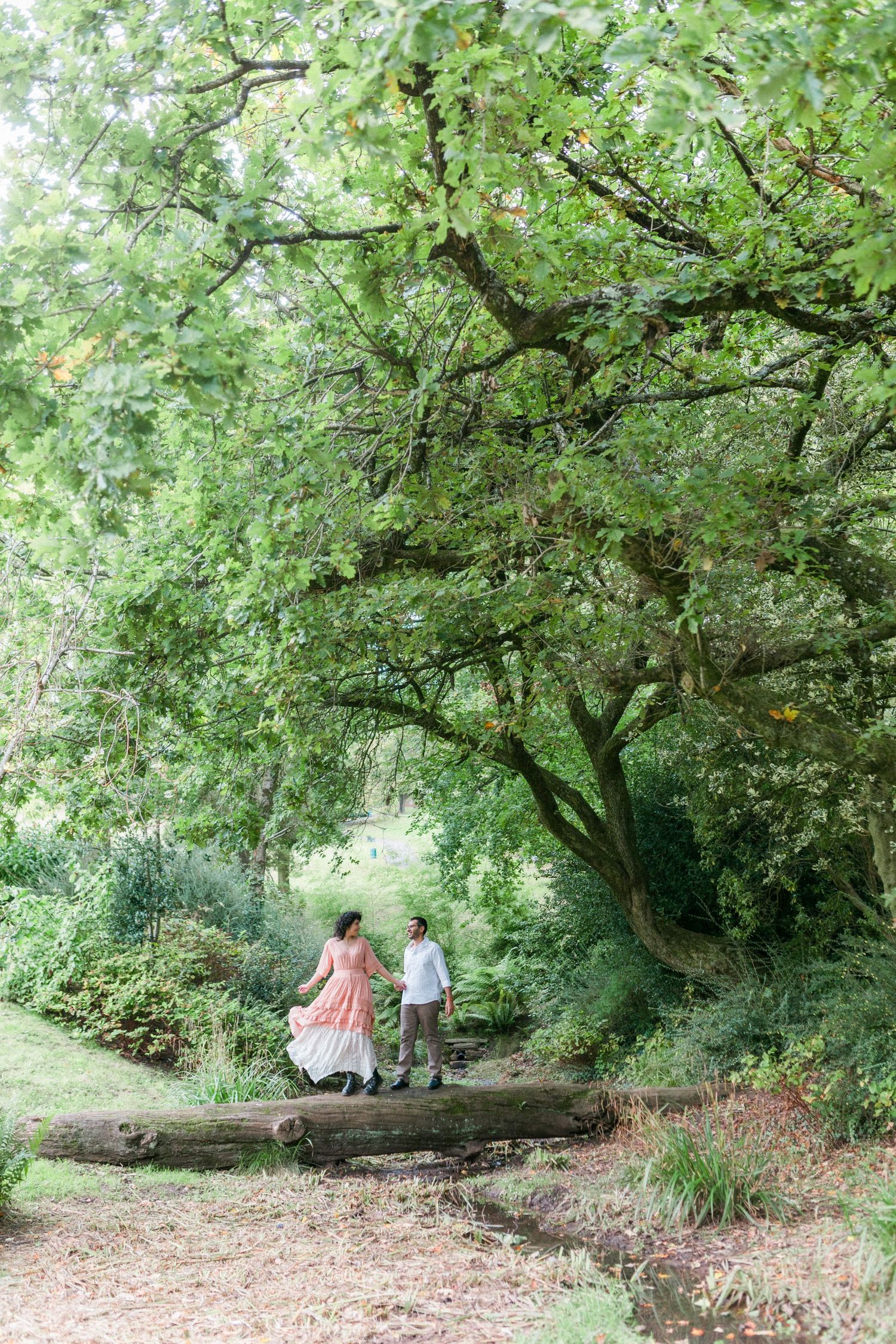 Two people enjoy a peaceful walk along a shaded forest path with lush green trees overhead and fallen logs on the ground.