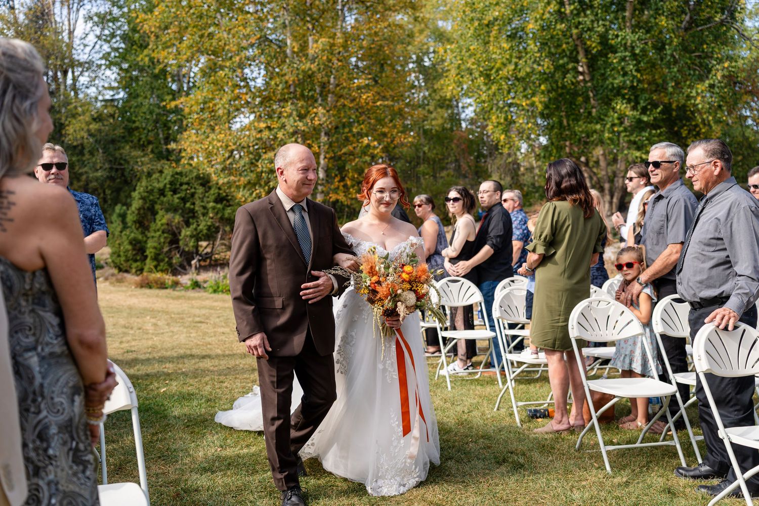 Wedding guests seated in white chairs on a lawn during an outdoor autumn ceremony with golden foliage in background.