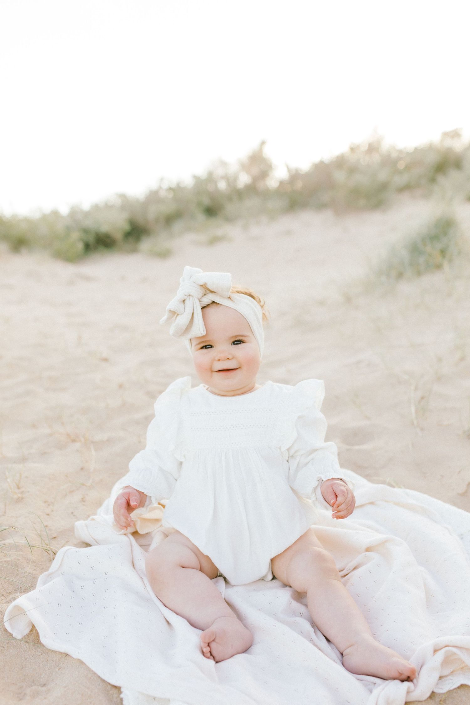 A baby in a white dress lays on a sandy beach blanket while smiling and playing.