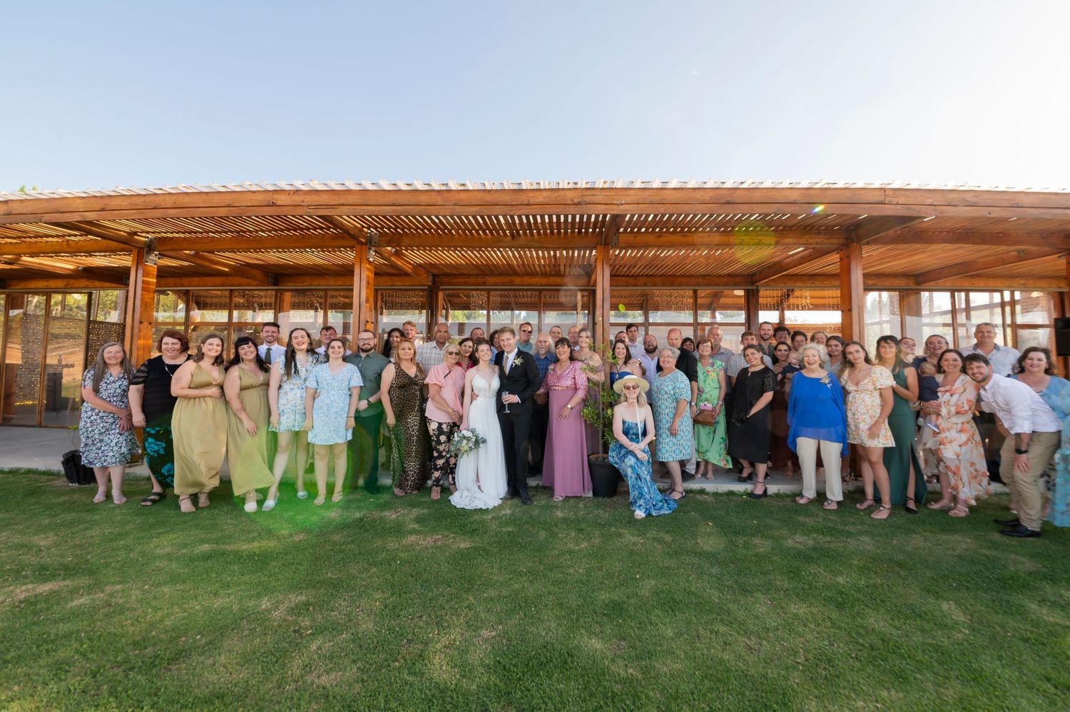 Large group of wedding guests gathered outdoors under wooden pergola structures during a summer celebration.