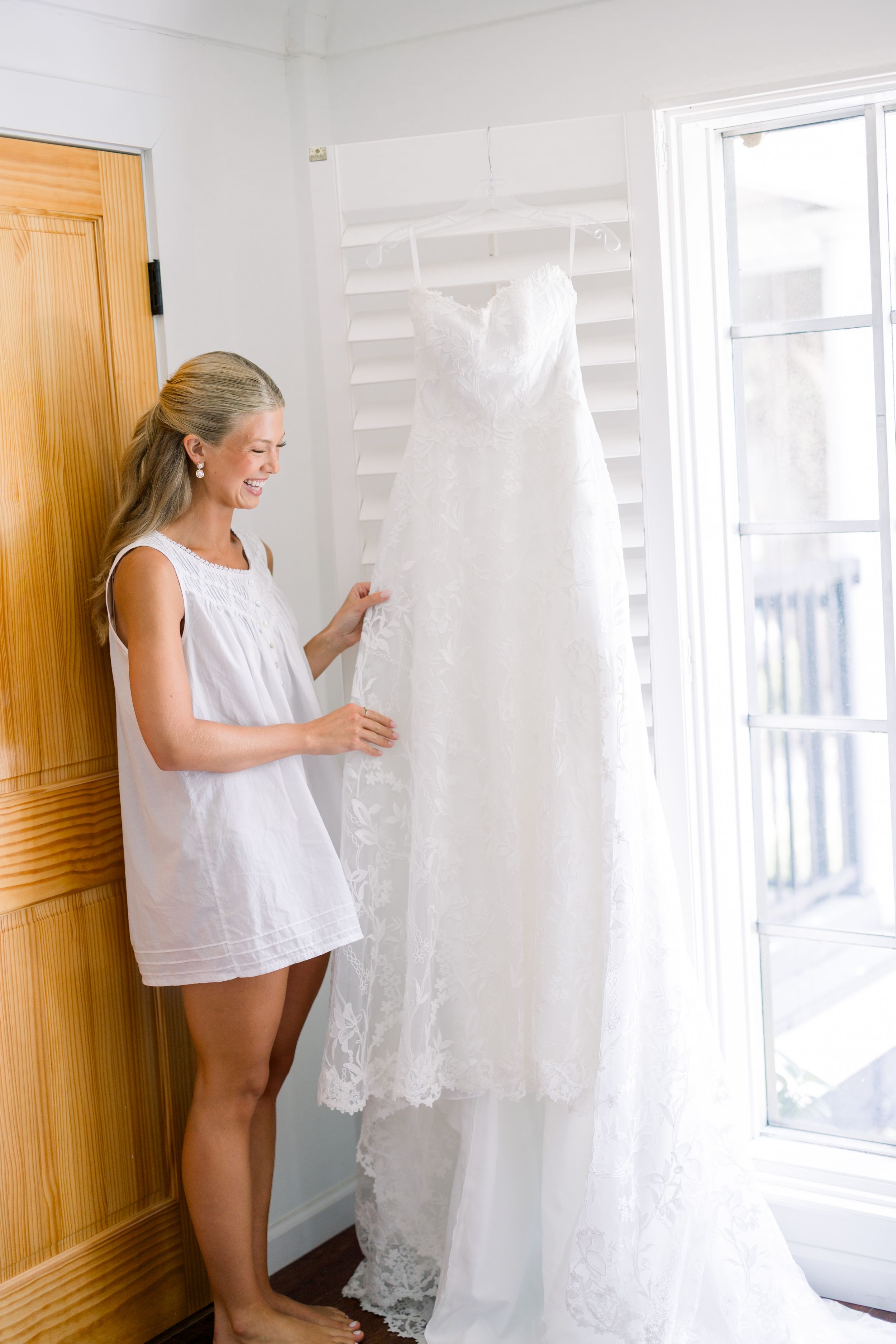 Person in white dress admiring wedding gown hanging by window in sunlit room.