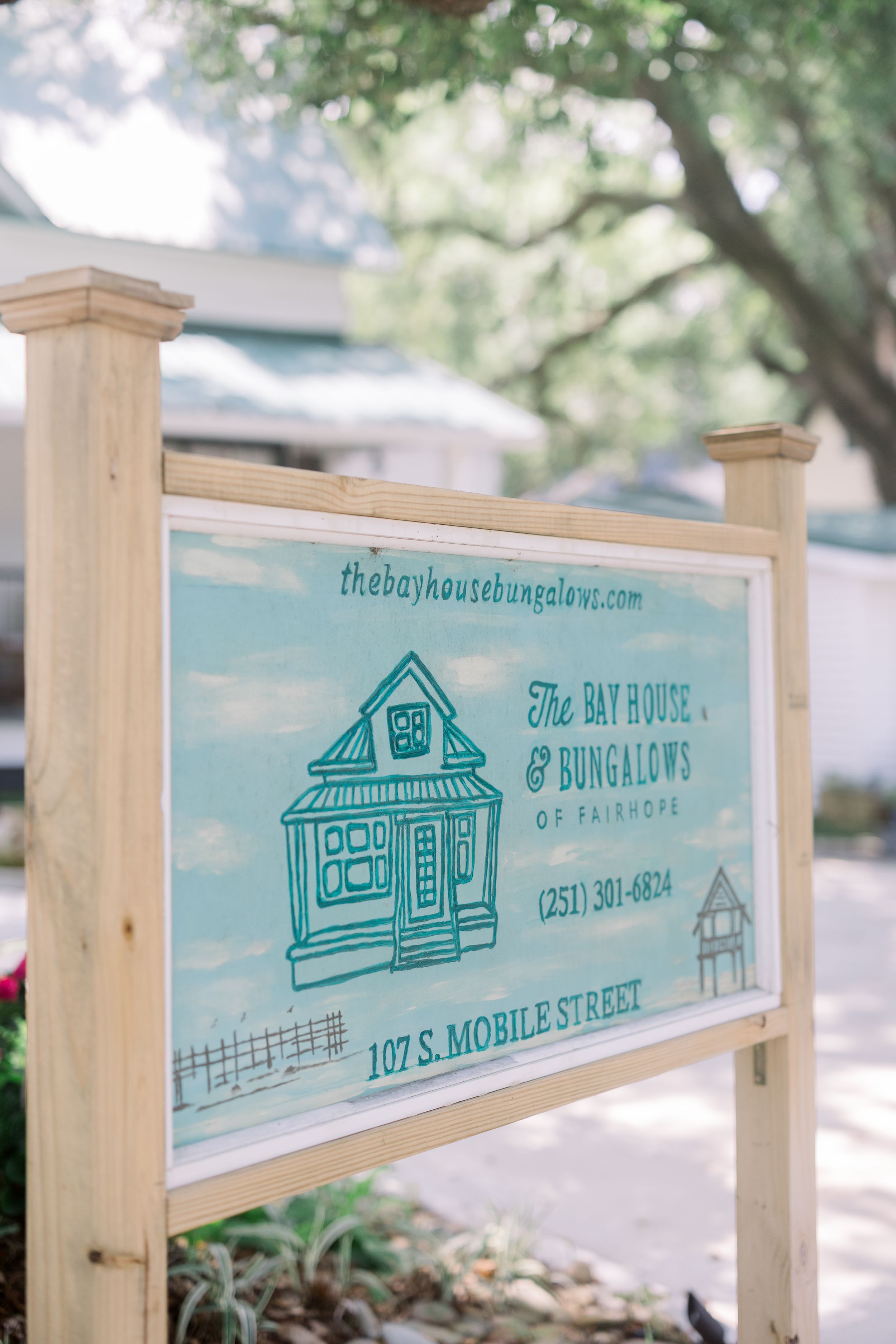 Wooden sign displaying 'The Bay House & Bungalows' with a cute cottage illustration in turquoise at 107 S Mobile Street.