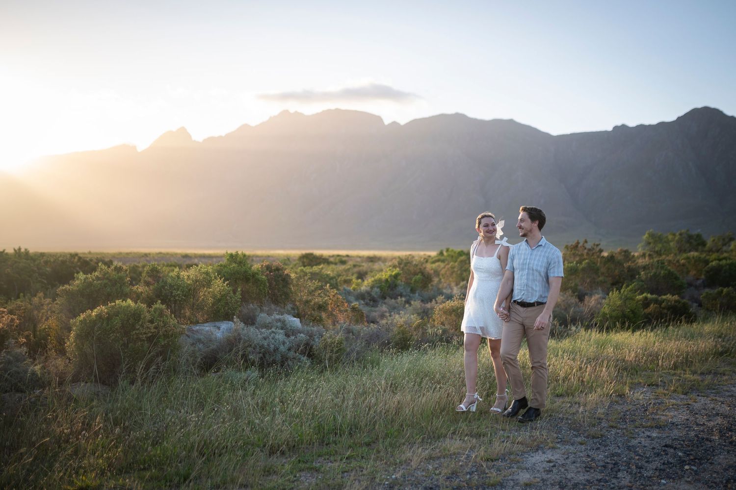 A couple walks along a mountain trail during golden hour with scenic mountain ranges in the background.