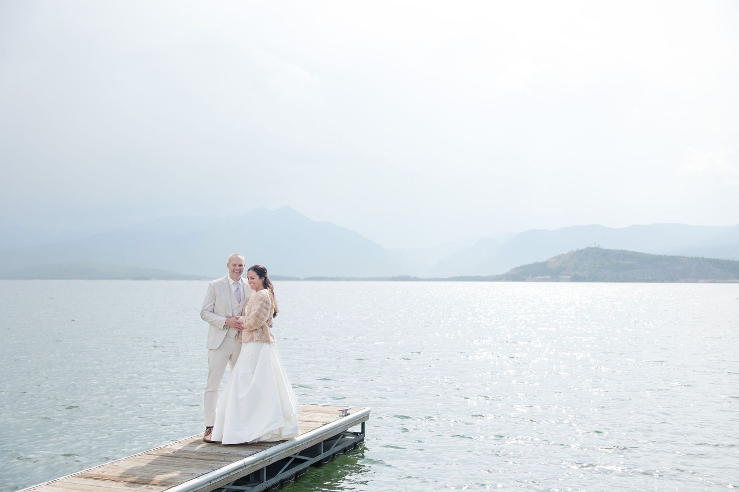 Couple in wedding attire embraces on a scenic pier overlooking misty mountains and water.